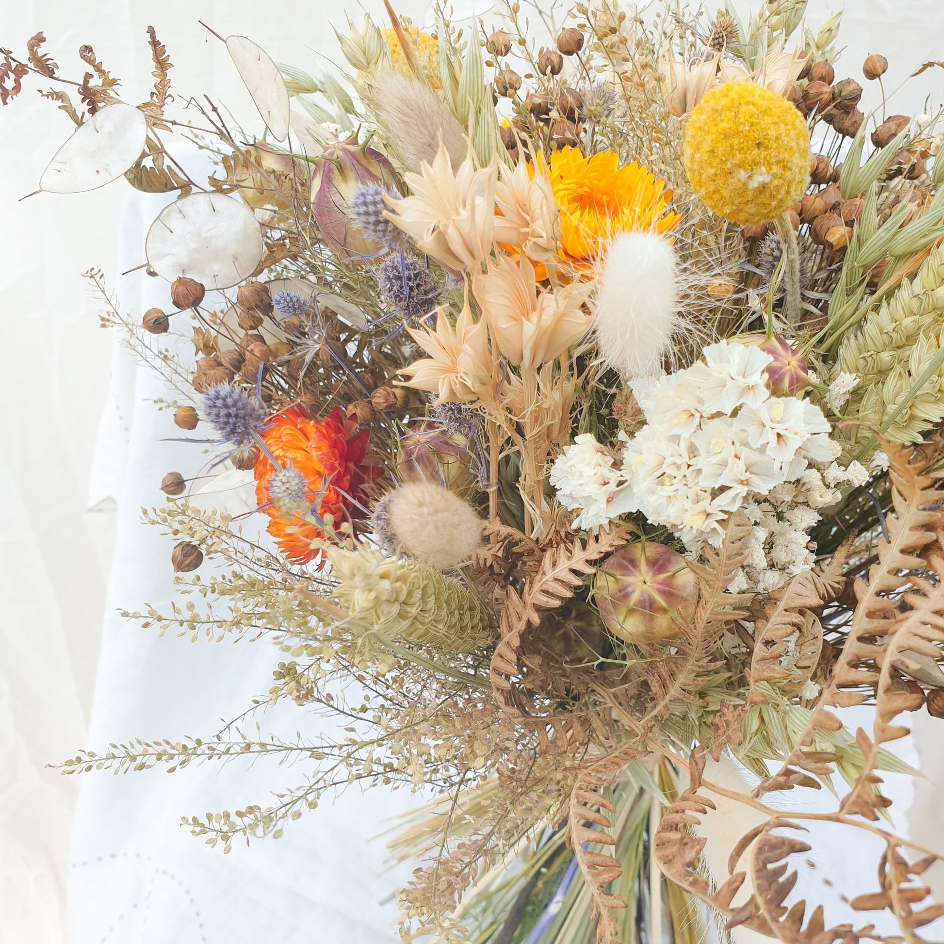 A close up of the Autumnal themed dried flower bouquet. You can see orange and yellow dried flowers, soft creamy white statice and silvery honesty. There are also small blue thistles and fluffy bunny tail grasses along with green and brown filler stems and natural Welsh bracken foliage.