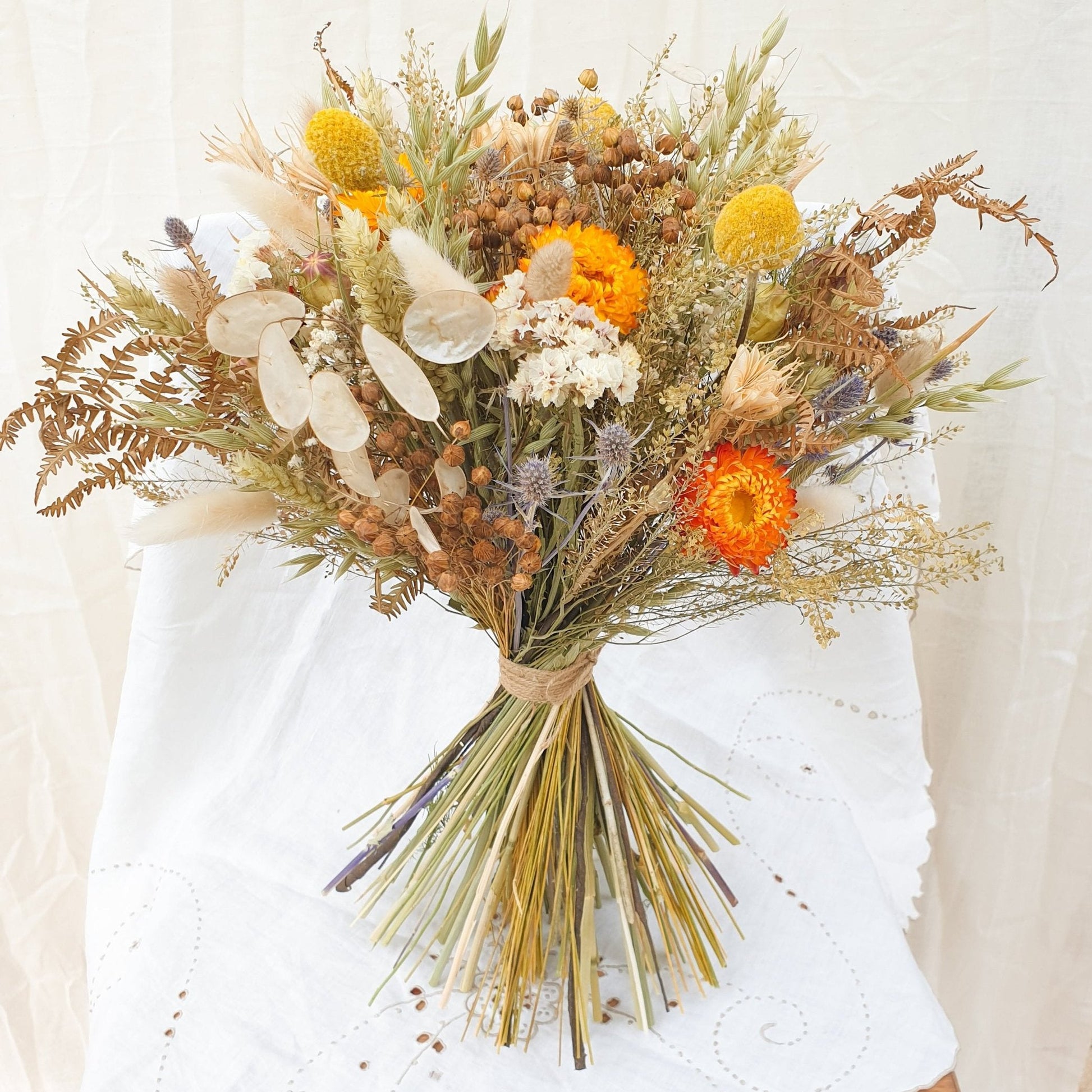 An Autumnal themed dried flower bouquet in a rounded handtie style. Orange and yellow dried flowers are accented with blue thistles, silvery honestly lunaria and fluffy bunny tail grasses. There are green and brown filler stems and natural Welsh bracken as foliage. The bouquet is informal, romantic and wild, as if it has been gathered together on a walk in the countryside. It is finished with a binding of rustic twine.