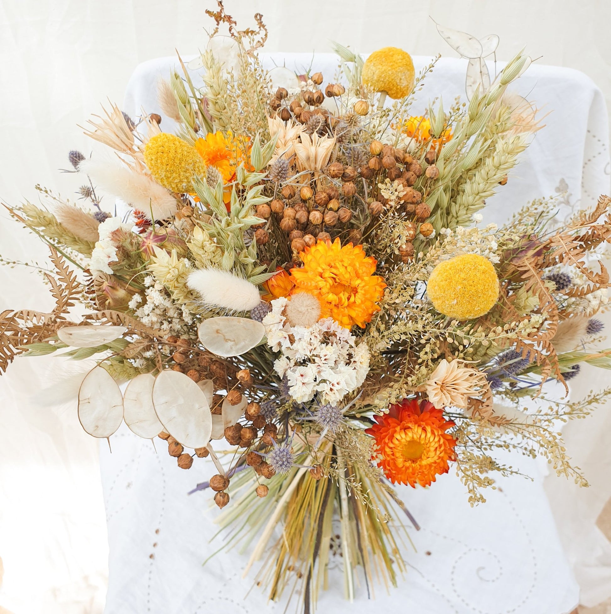 An Autumnal themed dried flower bouquet in a rounded handtie style. Orange and yellow dried flowers are accented with blue thistles, silvery honestly lunaria and fluffy bunny tail grasses. There are green and brown filler stems and natural Welsh bracken as foliage. The bouquet is informal, romantic and wild, as if it has been gathered together on a walk in the countryside. It is finished with a binding of rustic twine.