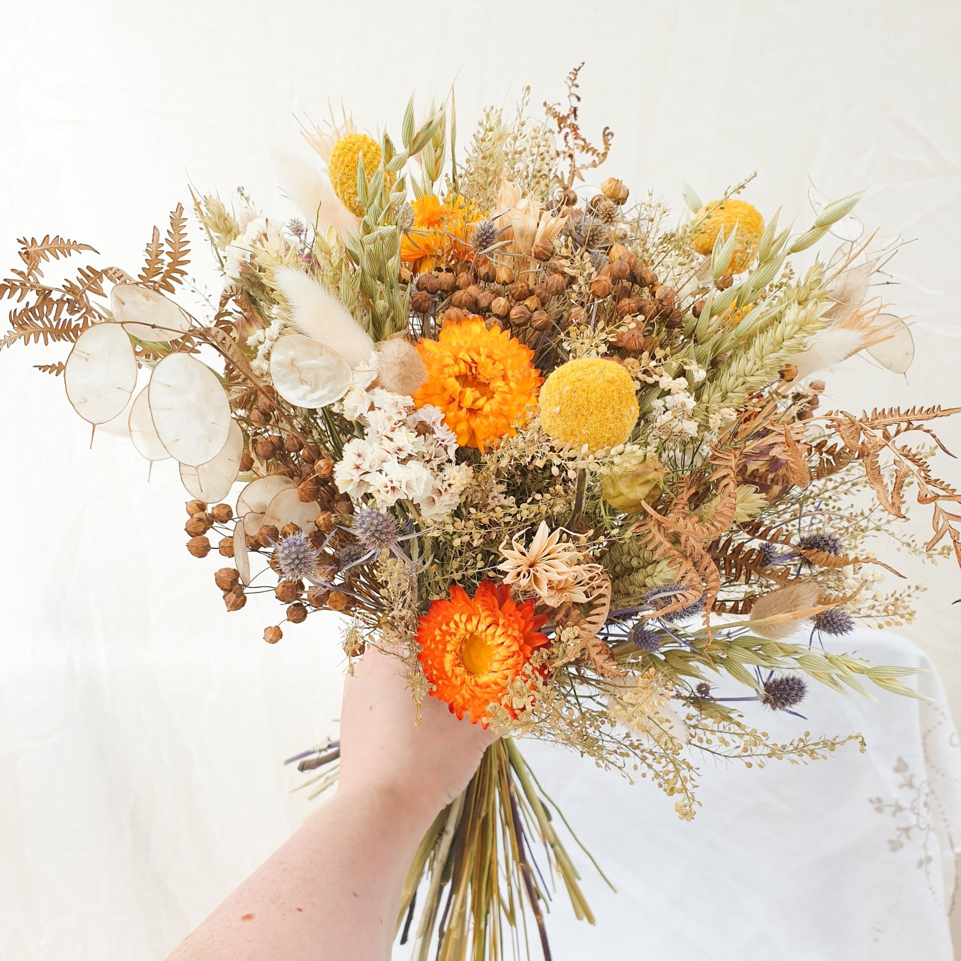 An Autumnal themed dried flower bouquet in a rounded handtie style is held in the hand, showing it is moderately large.