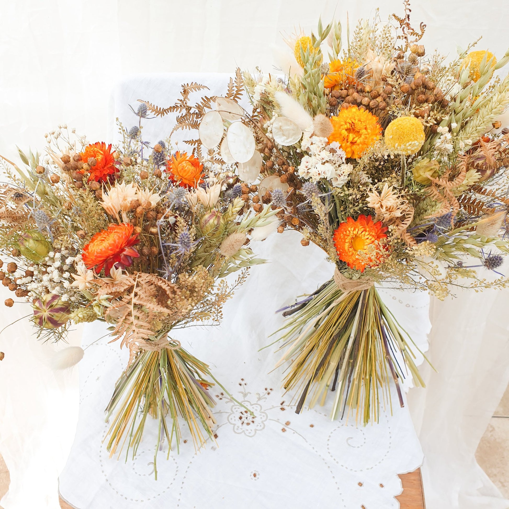 The two sizes of bouquet are shown next to each other. You can see the larger bouquet contains added yellow flowers and honesty, while the smaller bouquet focuses on the orange and blue flowers. The small version does not contain honesty. Both are shown tied with twine and have a rounded but informal wild look.