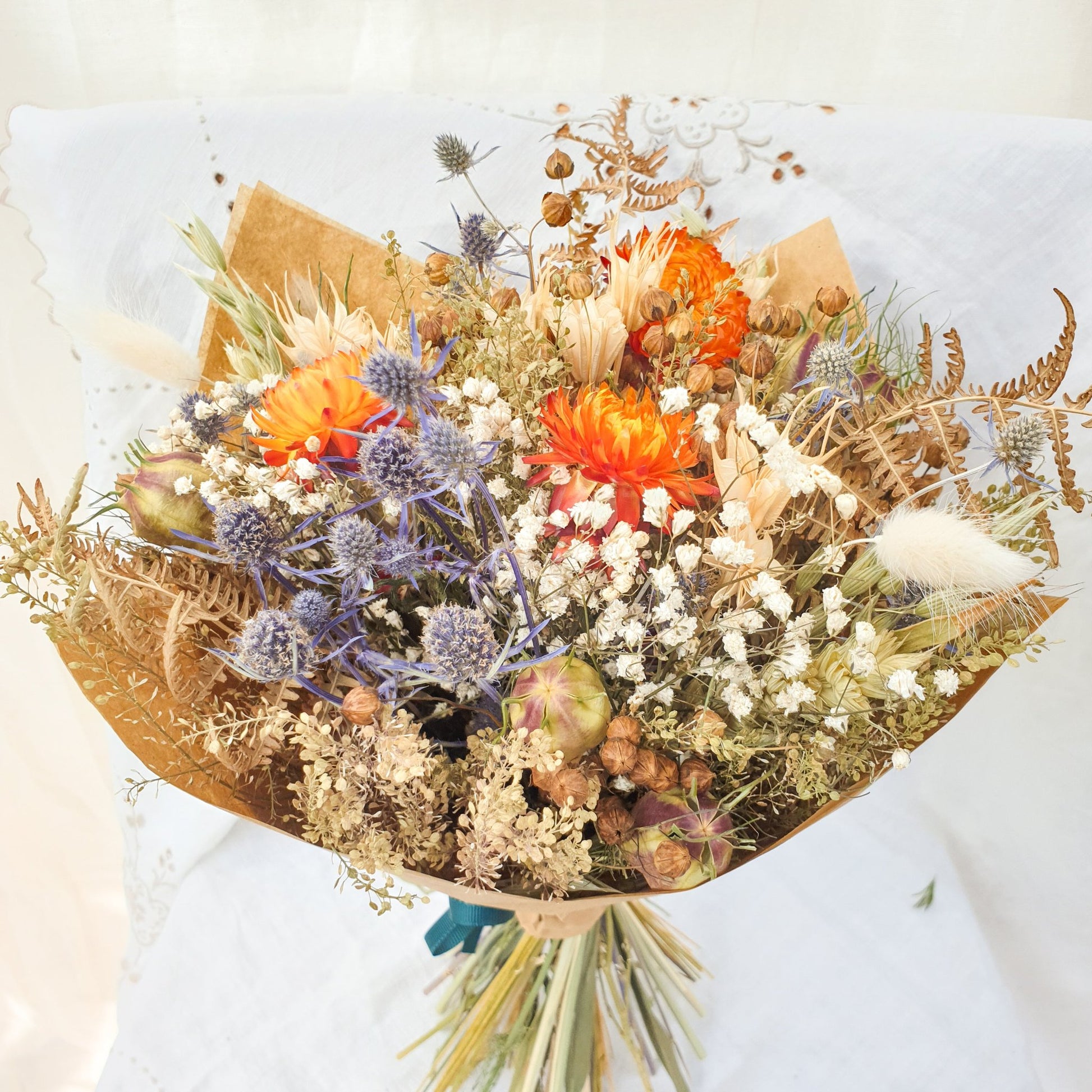 A close up of the smaller bouquet wrapped stylishly in brown paper. You can see it contrains orange dried flowers contrasted with small blue thistles and fluffy bunny tail grasses along with green and brown filler stems and natural Welsh bracken foliage.