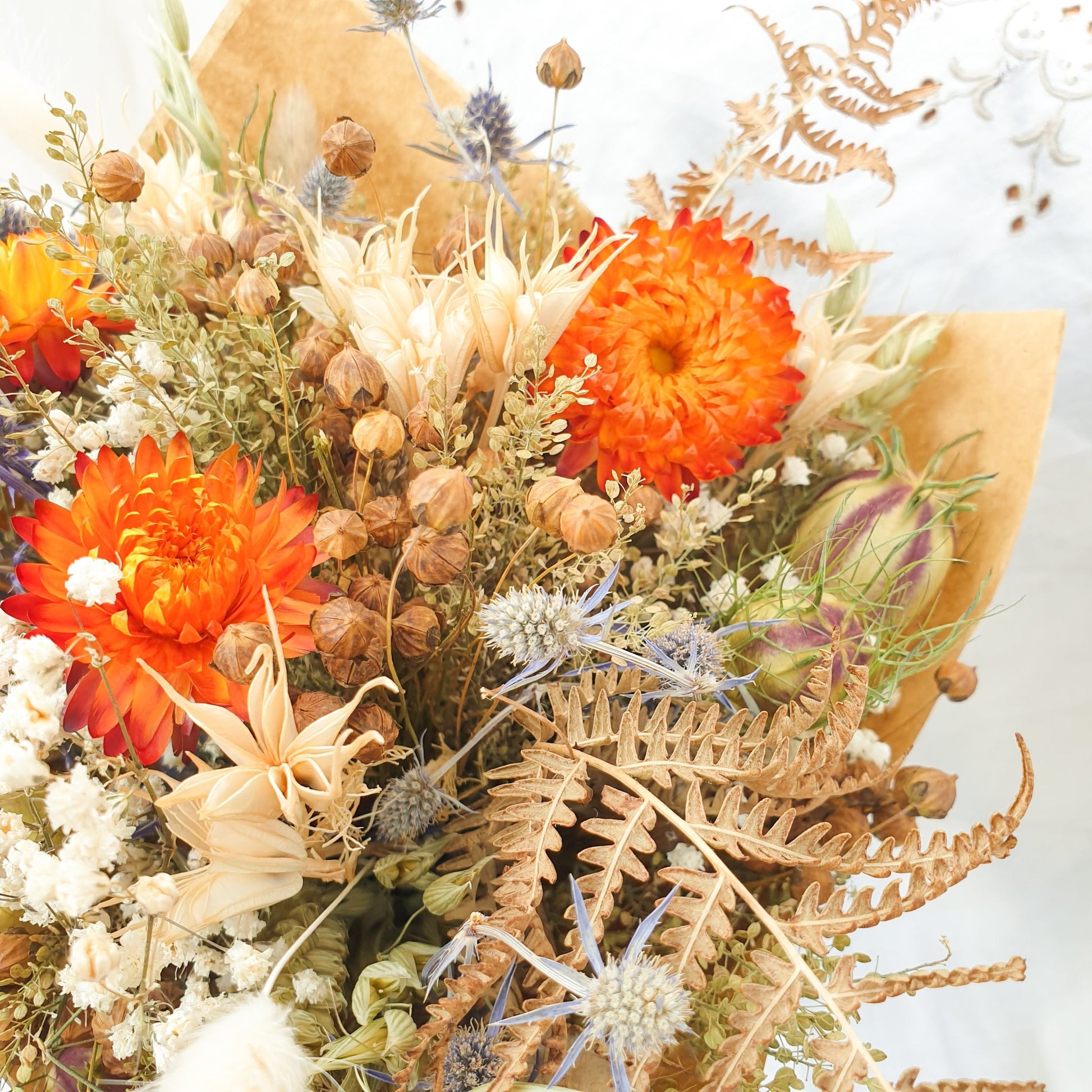 A close up of the smaller bouquet. You can see it contrains orange dried flowers contrasted with small blue thistles and fluffy bunny tail grasses along with green and brown filler stems and natural Welsh bracken foliage.