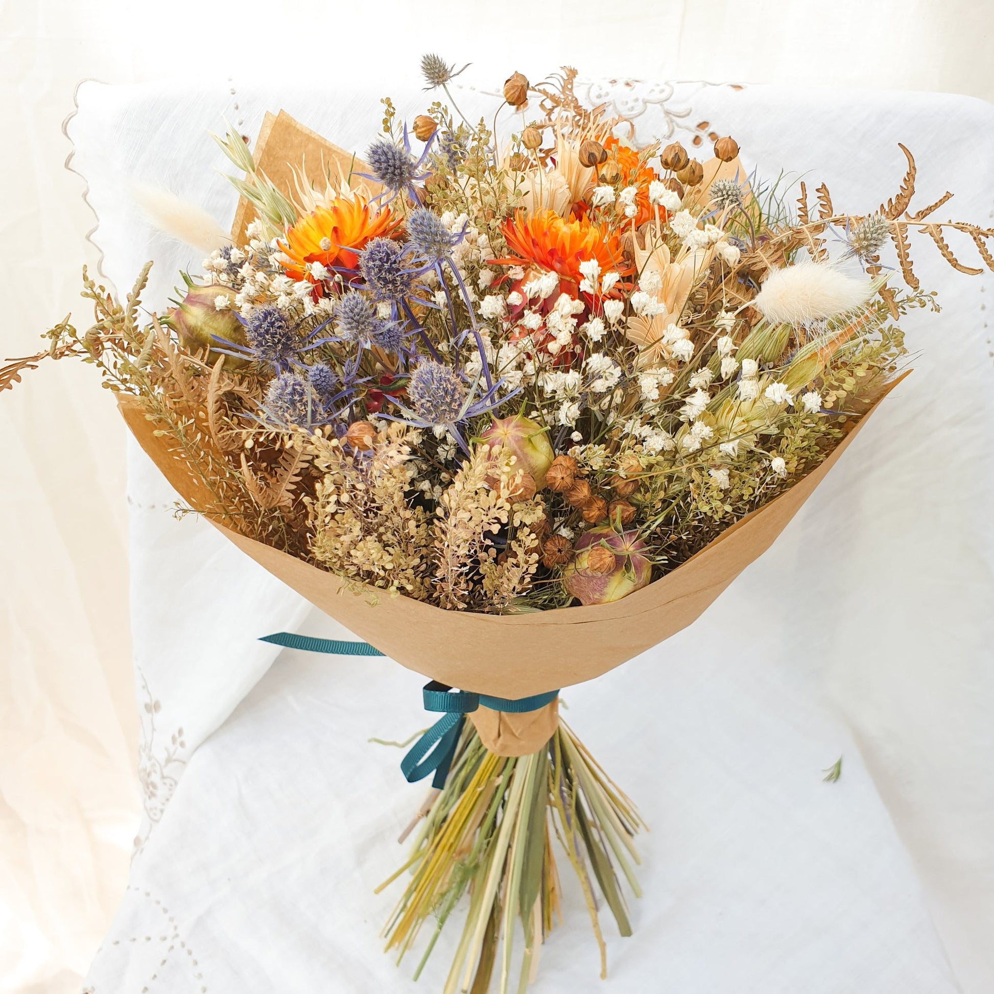 A close up of the smaller bouquet wrapped stylishly in brown paper held. You can see orange dried flowers contrasted with small blue thistles and fluffy bunny tail grasses along with green and brown filler stems and natural Welsh bracken foliage. It has a rounded yet informal and whimsical natural style.