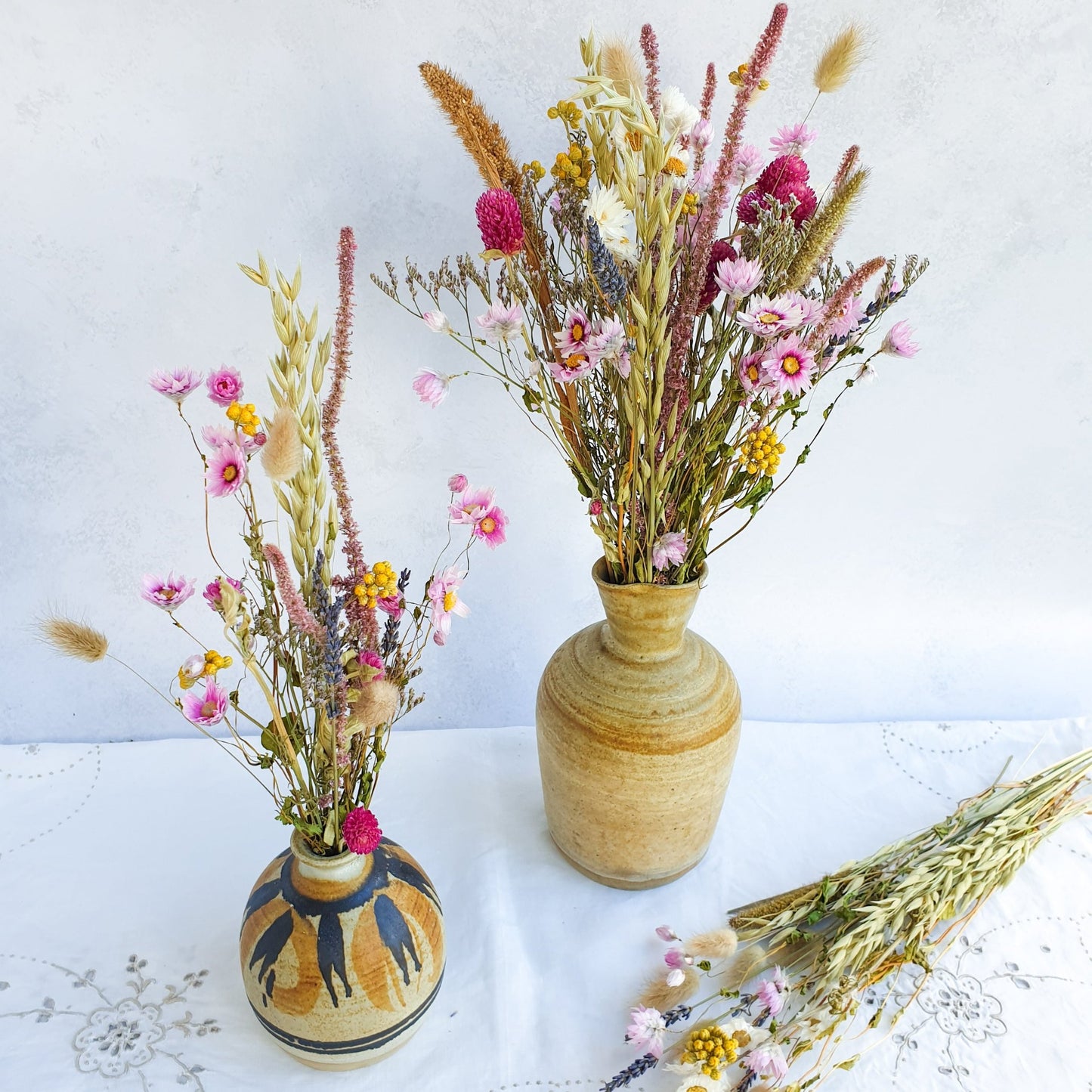 Dried Flower Mixed Box- Wildflower Fields by Lytton Rose Botanical. Two ceramic vases filled with vibrant dried flower arrangements. The smaller vase, decorated with a brushstroke pattern, holds a bouquet of pink and white flowers, including pink daisies and sea lavender, interspersed with dried grasses. The larger, textured vase displays a bountiful arrangement of pink and yellow flowers, including yellow lona and clovers, complemented by dried grasses and scented lavender.