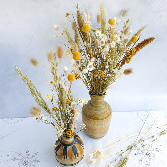 Dried Flower Mixed Box of Meadow Style Loose Stems- Summer Gold by Lytton Rose Botanical. A brown ceramic vase is filled with a vibrant bouquet of dried flowers. The bouquet features a mix of white daisies, yellow craspedia, and dried grasses, including lagurus fluffy bunny tail grass and wheat.