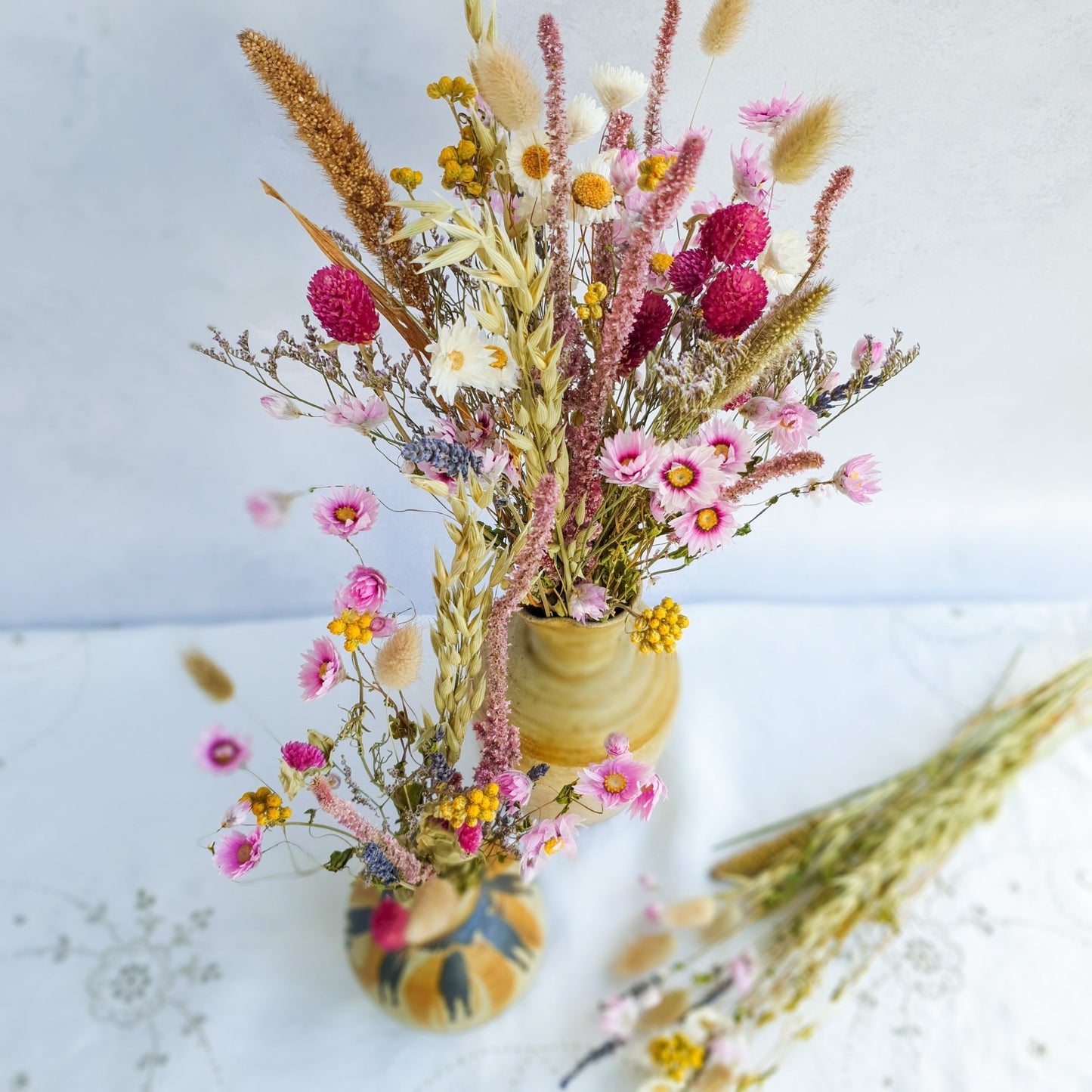 Dried Flower Mixed Box- Wildflower Fields by Lytton Rose Botanical. Two ceramic vases filled with vibrant dried flower arrangements. The smaller vase, decorated with a brushstroke pattern, holds a bouquet of pink and white flowers, including pink daisies and sea lavender, interspersed with dried grasses. The larger, textured vase displays a bountiful arrangement of pink and yellow flowers, including yellow lona and clovers, complemented by dried grasses and scented lavender.